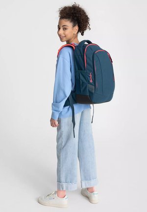 Young person with curly hair wearing a blue sweatshirt, light jeans, and white sneakers, carrying a large blue backpack with pink accents.