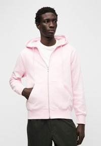 Jasnoróżowa bluza z kapturem na zamek, wykonana z miękkiego materiału, z przednią kieszenią, kapturem ze sznurkiem i małym białym logo na piersi.