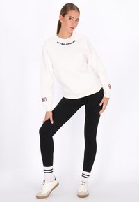 Weißes Sweatshirt mit schwarzem Text, schwarze Leggings und weiße Turnschuhe. Verfügt über gerippte Bündchen und ein Logo am Ärmel. Einfaches, sportliches Design.