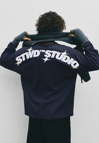 Persona con capelli ricci che indossa una camicia blu navy con la scritta "STWD STUDIO" sulla schiena, tenendo una sciarpa scura dietro al collo.