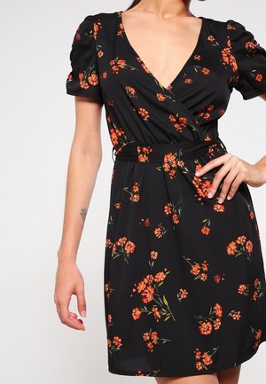 Černé zavinovací šaty s krátkými nabíranými rukávy a oranžovým květinovým vzorem, oblečené na osobě, která má jednu ruku na bok a druhou volně spuštěnou.