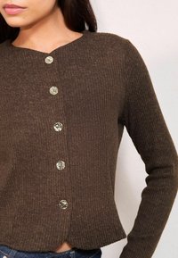 Hnedý rebrovaný kardigán s okrúhlym výstrihom, ktorý má päť lesklých gombíkov na prednej strane. Spodný lem je mierne vlnitý, čo pridáva na detailoch.