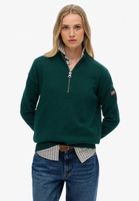 Groene gebreide trui met een ritskraag, voorzien van ribgebreide manchetten en zoom, gedragen over een blauw-wit gestreept shirt, gecombineerd met blauwe jeans.