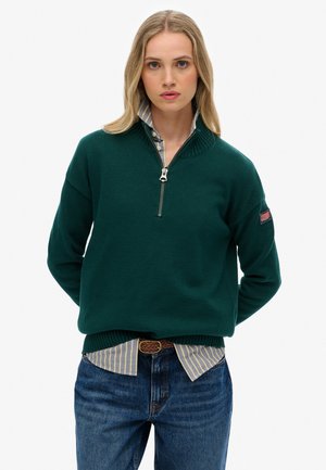 Groene gebreide trui met een ritskraag, voorzien van ribgebreide manchetten en zoom, gedragen over een blauw-wit gestreept shirt, gecombineerd met blauwe jeans.