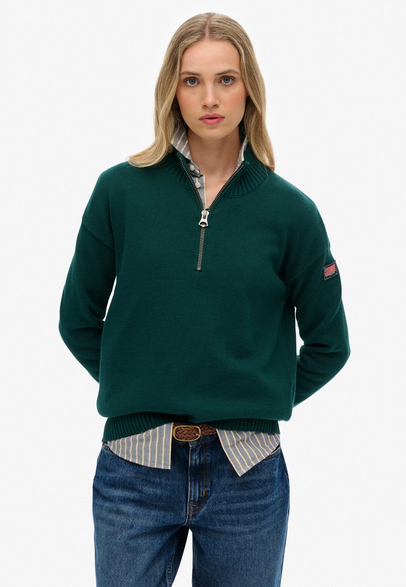 Groene gebreide trui met een ritskraag, voorzien van ribgebreide manchetten en zoom, gedragen over een blauw-wit gestreept shirt, gecombineerd met blauwe jeans.