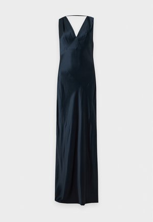 Une femme porte une robe longue jusqu’au sol, bleu marine foncé, sans manches et à col en V, avec des chaussures beiges, sur un fond clair uni.