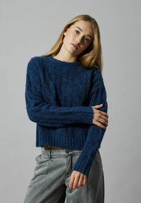 Niebieski sweter z dzianiny z ribbowanym dołem i mankietami, z teksturowanym wzorem. Połączony z jasnoszarymi spodniami. Prosty, skrócony krój.