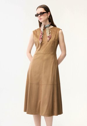 Vrouw die een mouwloze lichtbruine midi-jurk draagt, met een sjaal met patroon om de hals geknoopt, kleine rechthoekige zonnebril en gouden oorringen, staand met haar handen achter haar rug.