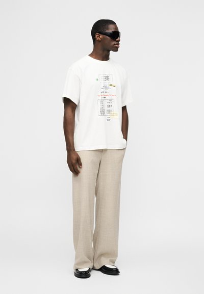 Άνδρας με μαύρα μεγάλα γυαλιά ηλίου, λευκό T-shirt με γραφικό σχέδιο, μπεζ παντελόνι φαρδύ, και ασπρόμαυρα loafers, στέκεται μπροστά σε μονόχρωμο φόντο.