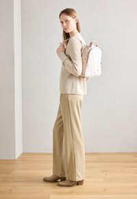 Zaino con motivo floreale nei toni del rosa e del beige chiaro. Maglione beige a coste e pantaloni beige a gamba larga. Scarpe con tacco marroni. Sfondo in legno.