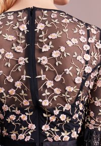 Haut en tissu noir transparent avec une broderie florale rose intricatement réalisée, des accents métalliques et une fermeture éclair au dos pour un look détaillé et texturé.