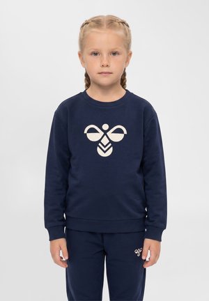 Młoda dziewczyna z blond plecionymi włosami, ubrana w granatową bluzę i spodnie z białym logo pszczoły na piersi i nodze.