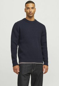 Navyblauer, gerippter Strickpullover mit langen Ärmeln, rundem Halsausschnitt und enganliegendem Saum; über einem grauen Hemd getragen, kombiniert mit dunklen Jeans.