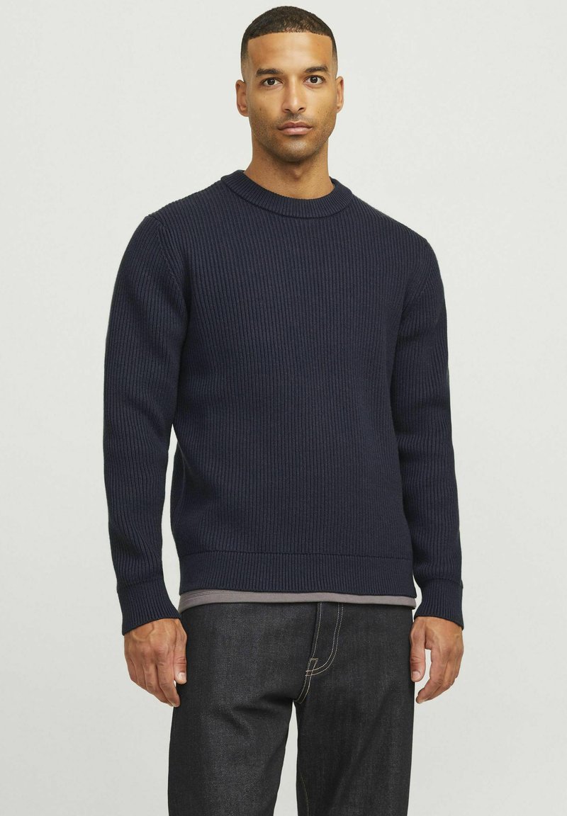 Navyblauer, gerippter Strickpullover mit langen Ärmeln, rundem Halsausschnitt und enganliegendem Saum; über einem grauen Hemd getragen, kombiniert mit dunklen Jeans.
