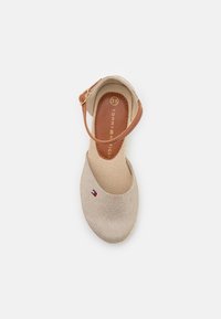 Beżowe płócienne buty z brązowym paskiem na kostkę, okrągłym noskiem i teksturowaną powierzchnią. Zawierają małe logo w pobliżu obszaru palców.