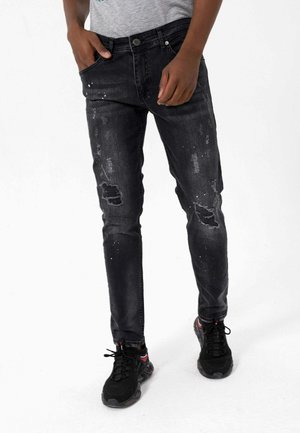 Personne portant un jean noir déchiré avec des éclaboussures de peinture et des baskets noires, debout avec une main dans la poche.