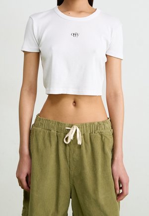 Femme portant un haut court blanc à manches courtes avec un petit logo noir et un pantalon ample vert olive avec cordon, sur un fond uni.