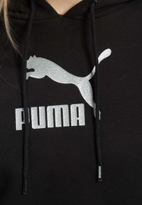 Čierna mikina s kapucňou, na prednej strane s veľkým, trblietavým strieborným logom Puma a textom "PUMA". Vyrobená z jemného materiálu s nastaviteľnými sťahovacími šnúrkami.