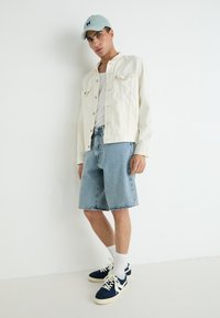 Veste en denim blanc, casquette bleu clair, t-shirt blanc, shorts en denim bleu et baskets navy avec des accents blancs. Tenue simple et décontractée.