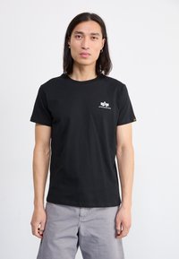Schwarzes Baumwoll-T-Shirt mit Rundhalsausschnitt, das ein weißes Logo auf der linken Brust hat. Das Modell trägt hellgraue Hosen.