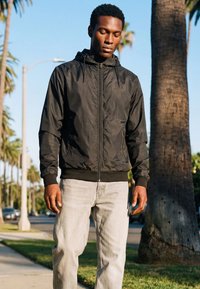 Man draagt een zwart jack met rits en lichte spijkerbroek, staat buiten bij een palmboom op een zonnige dag.