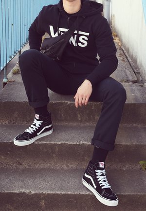 Person trägt schwarzen Vans-Hoodie, schwarze Hose, schwarz-weiße Vans-Sneaker und schwarze Umhängetasche, sitzt auf einer Außentreppe aus Beton.
