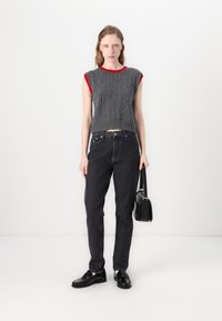 Crop top en tricot gris avec des finitions rouges, associé à un jean noir taille haute. Le mannequin tient un petit sac noir et porte des chaussures noires.