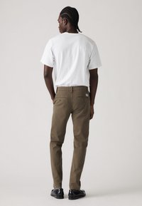 Brown, straight-leg pants made of cotton with back pockets, worn with a white t-shirt. Features a label on the waistband. Black shoes.
