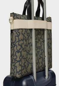 Bolsa de tote con patrón negro y beige, con doble asa, sujeta a una maleta azul con ruedas mediante una correa en contraste. Acabado de tela texturizada.