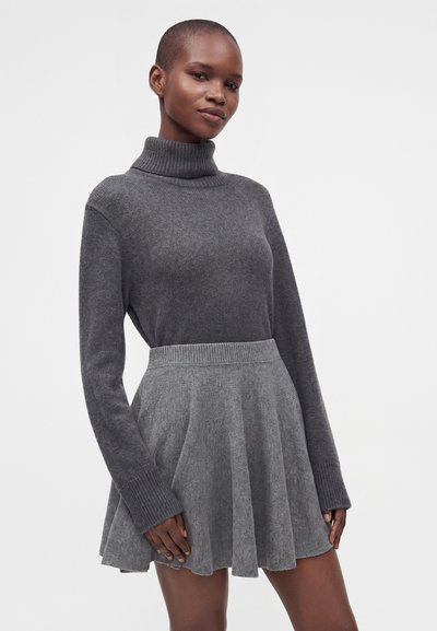 Femme portant un pull à col roulé gris foncé et une jupe tricotée gris clair sur un fond blanc uni.