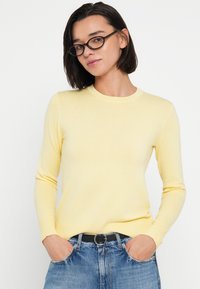 Κίτρινο πουλόβερ με ριμπ πλέξη, μακρυά μανίκια και στρογγυλή λαιμόκοψη, συνδυασμένο με μπλε τζιν και μαύρη ζώνη.