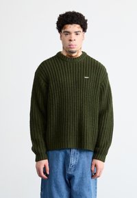 Πλεκτό πράσινο πουλόβερ με ριγωτή υφή και στρογγυλή λαιμόκοψη, με μικρό λευκό λογότυπο "OBEY". Συνδυασμένο με μπλε τζην παντελόνι.