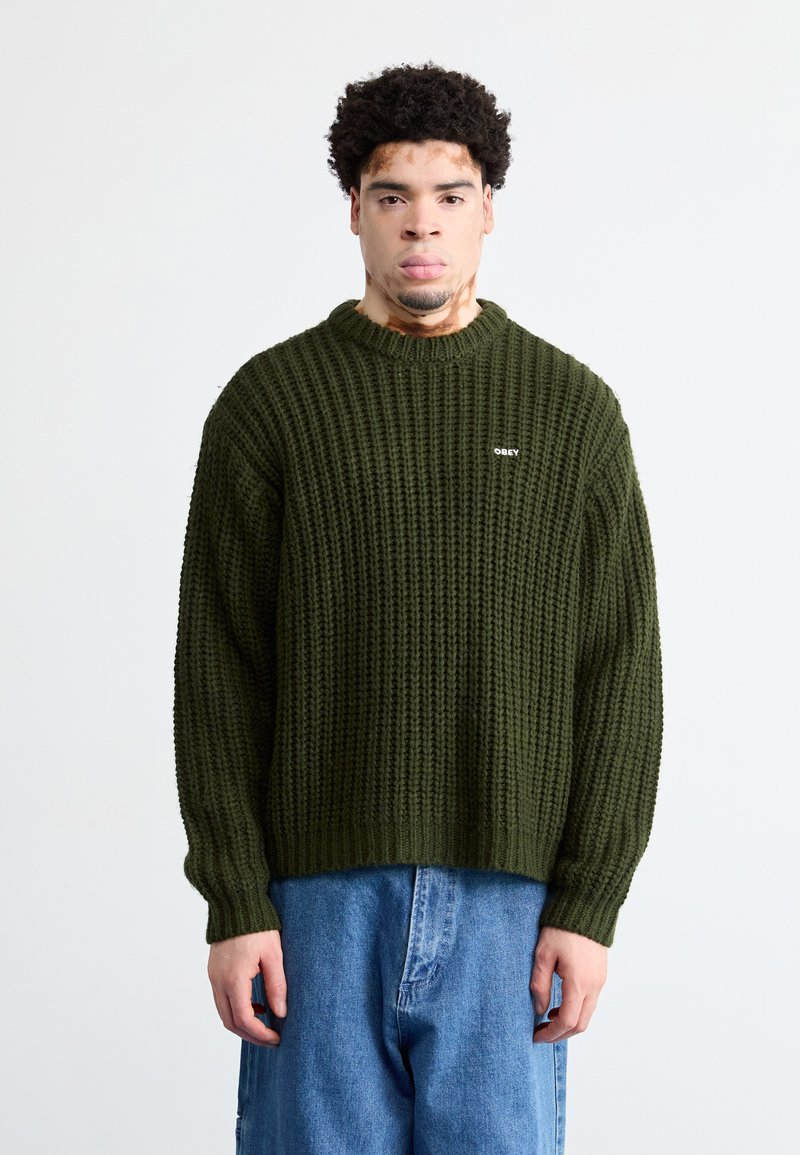 Πλεκτό πράσινο πουλόβερ με ριγωτή υφή και στρογγυλή λαιμόκοψη, με μικρό λευκό λογότυπο "OBEY". Συνδυασμένο με μπλε τζην παντελόνι.