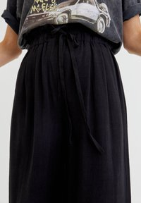 Osoba ubrana w czarne spodnie z troczkami oraz czarną koszulkę z graficznym nadrukiem z podwiniętymi rękawami, przedstawiającą samochód w stylu vintage oraz tekst.