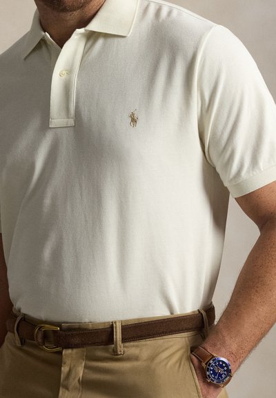 Homme portant un polo blanc, un pantalon beige, une ceinture en cuir marron et une montre marron avec un cadran bleu au poignet gauche.