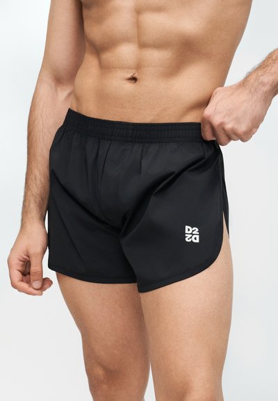 Ανδρικός κορμός και πάνω μέρος ποδιών που φορούν μαύρα αθλητικά σορτς με λευκό λογότυπο "DS2", χέρι που ρυθμίζει τη ζώνη σε απλό ανοιχτό φόντο.