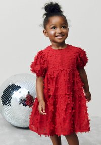 Rotes Kleid mit mehrlagigem Design, verziert mit floralen Applikationen und Puffärmeln. Glatte Textur, getragen von einem Kind neben einer spiegelnden Discokugel.