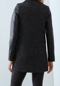 Černý rybí kost vlněný kabát s koženými rukávy, středně dlouhý střih, bezšvový zadní díl a kulatý límec, hladká textura a minimální kovové doplňky.