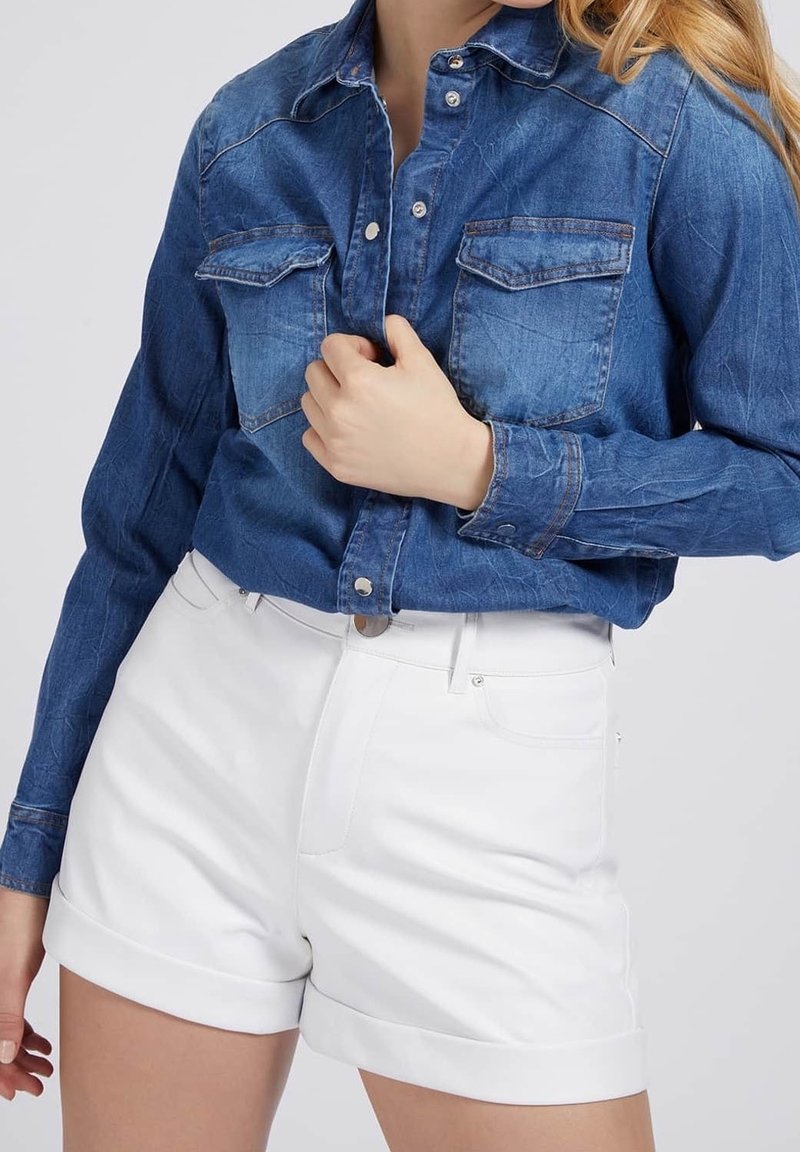 Camicia di denim blu medio con chiusura a bottoni e due tasche sul petto, abbinata a pantaloni corti bianchi con risvolto, dotati di parte anteriore piatta e passanti per la cintura.