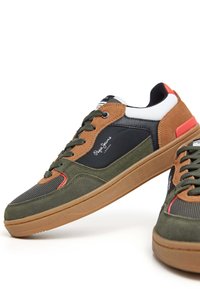 Zapatillas de ante verdes y marrones con paneles de malla negra, acentos rojos y suelas de goma marrones, que presentan cordones redondos y un logo en el lateral.