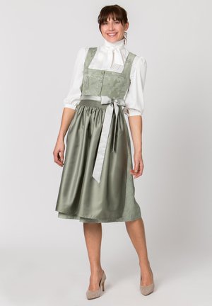Frau lächelt, trägt ein salbeigrünes traditionelles Kleid mit besticktem Mieder, passender Schürze, die an der Taille gebunden ist, weiße Bluse und beige Absätze.