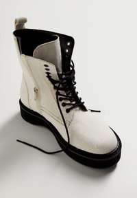 Botín de tobillo de piel de ternera blanca con acentos negros y suela gruesa. Cuenta con cordones en la parte delantera y cremallera lateral para un fácil acceso.