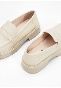 Mocassins beiges en cuir lisse, avec une semelle basse, une forme arrondie et des détails de couture subtils sur le dessus.