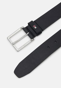 Cintura in pelle nera con fibbia rettangolare in metallo argentato, marchiata "TOMMY HILFIGER." Presenta una texture liscia e fori distribuiti uniformemente.