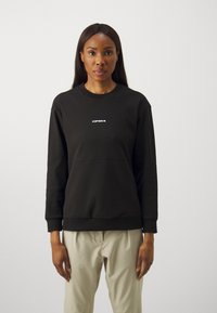 Black sweatshirt with ribbed texture, crew neck, and a visible logo in white. Casual fit and long sleeves, paired with light-colored trousers.