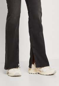 Zapatillas blancas con acentos texturizados y jeans negros de campana con aberturas en el dobladillo, sobre un fondo claro.