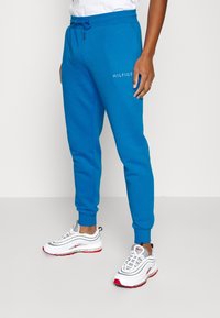 Blue sweatpants with elastic waistband and drawstring, featuring the logo "HILFIGER" in white on the side. Paired with white and red sneakers.