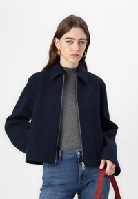 Giacca di lana blu navy con zip frontale e maniche larghe, dotata di colletto e tessuto texture. Abbinata a una maglia grigia e jeans blu.