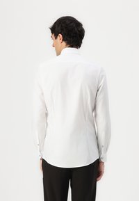 White button-up shirt with a tailored fit, featuring long sleeves, curved hem, and blue accents on the cuffs. Smooth fabric texture.