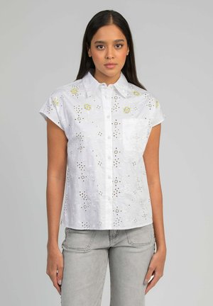 Femme portant une chemise blanche à manches courtes boutonnée avec broderies florales et un jean gris clair, debout devant un fond uni.